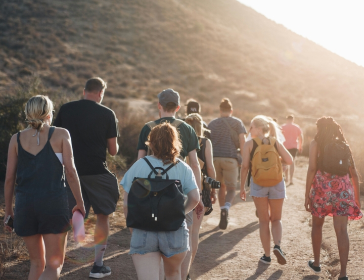 Groep reizigers wandelt met rugzakken over een zonnig pad door een heuvelachtig landschap.