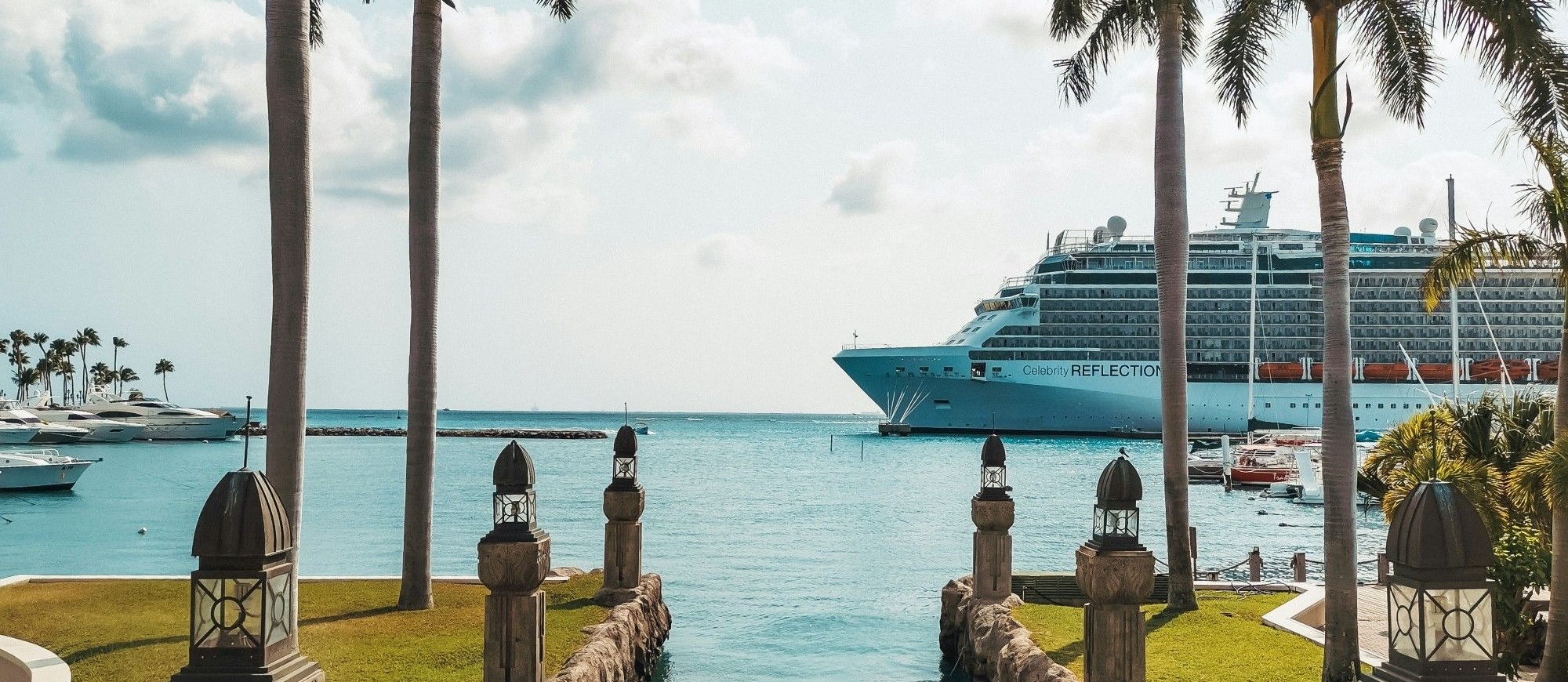 Tropische waterpartij met palmbomen en uitzicht op zee, met een groot cruiseschip op de achtergrond.